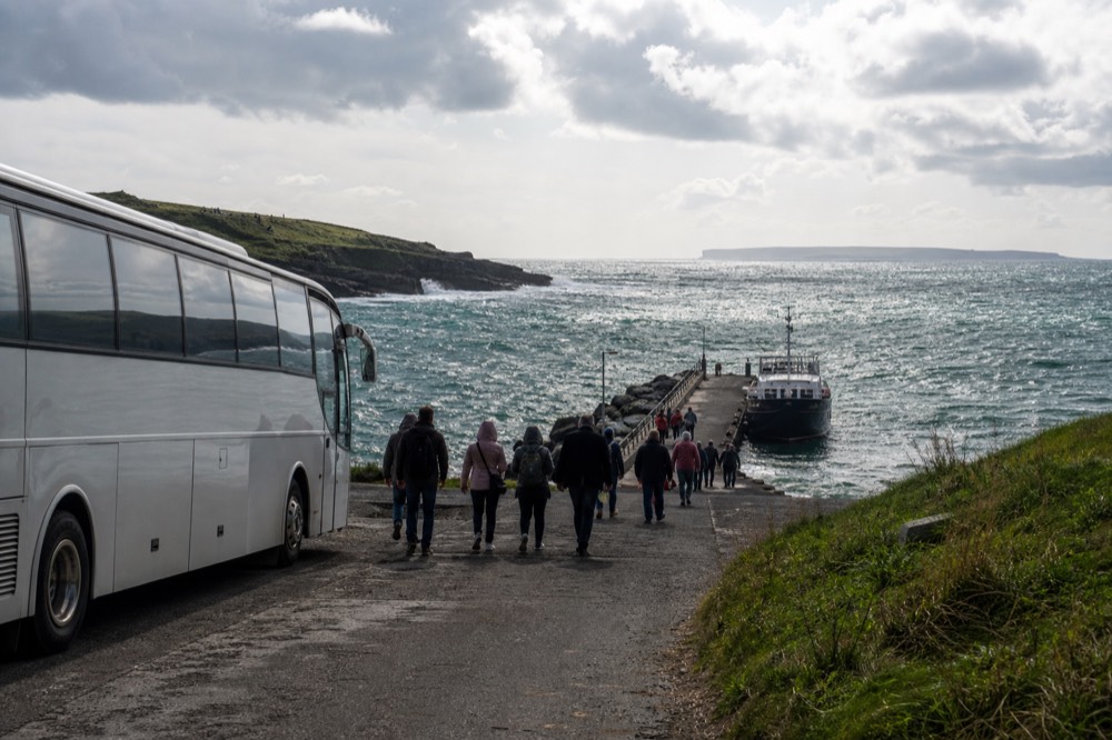 A tour coach parked beside Doolin harbour with passengers walking toward a waiting ferry, green hills behind and the Aran Islands low on the horizon.