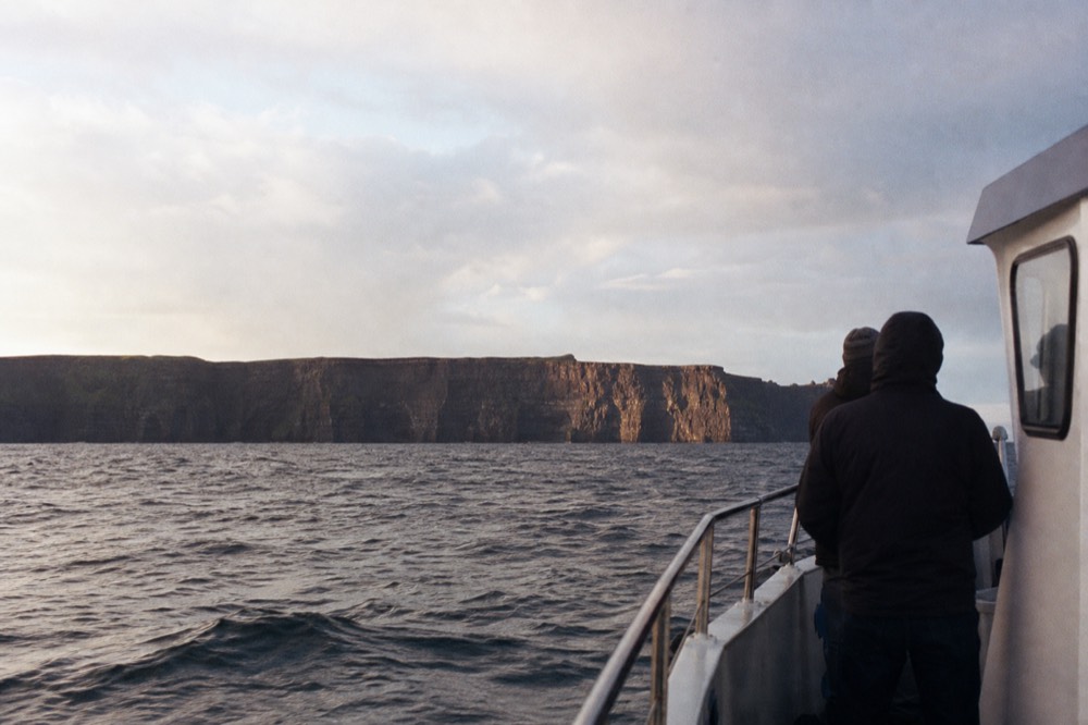 The full dark face of the Cliffs of Moher seen from a boat well out on the water, the wall small in the distance across grey Atlantic swell.