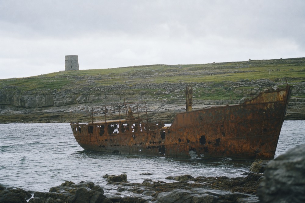 The rusting hull of the Plassey shipwreck on the rocky shore of Inis Oirr, a small stone tower and green fields rising behind it under a bright Atlantic sky.