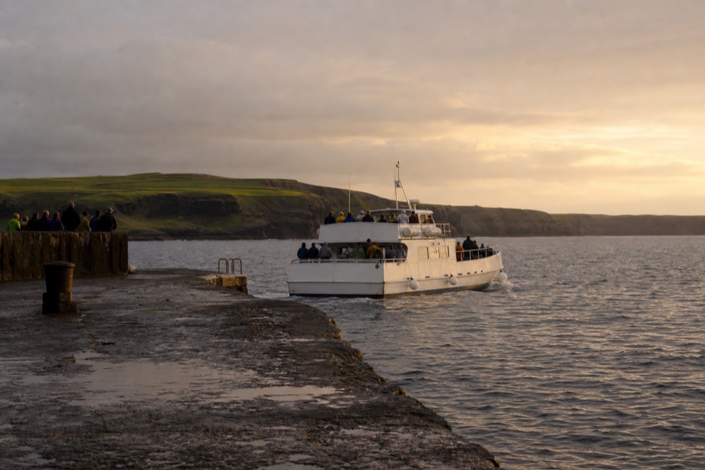 A white cliff-cruise boat pulling away from the stone jetty at Doolin Pier, green Clare hills behind it and open grey water ahead.