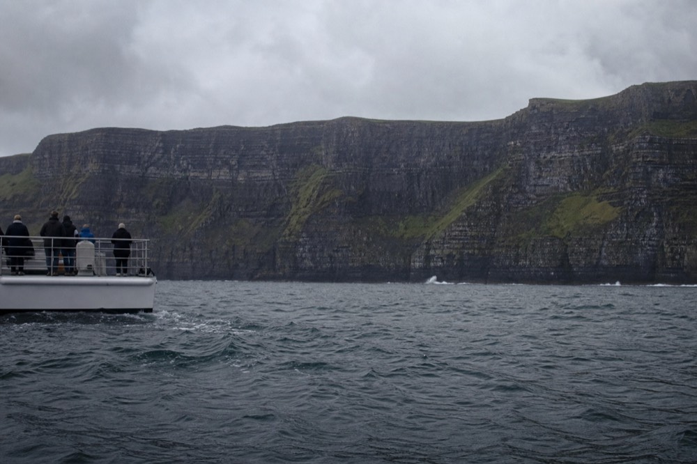 The full 214-metre face of the Cliffs of Moher seen from sea level, horizontal bands of dark rock streaked with green, a thin line of white surf at the base.