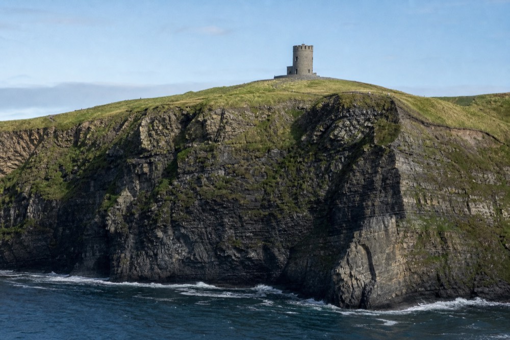 O'Brien's Tower, a round stone tower, standing on the green clifftop high above, seen from a boat far below with the cliff face falling away beneath it.