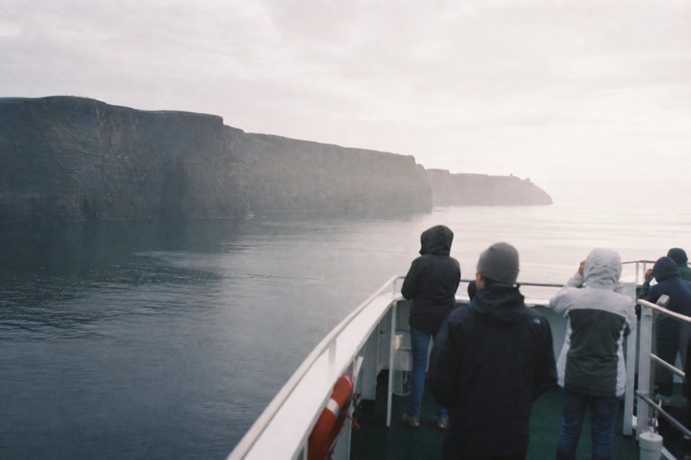 Passengers along the open deck of a returning cliff-cruise boat, some looking back at the Cliffs of Moher receding behind them in soft late-afternoon light.