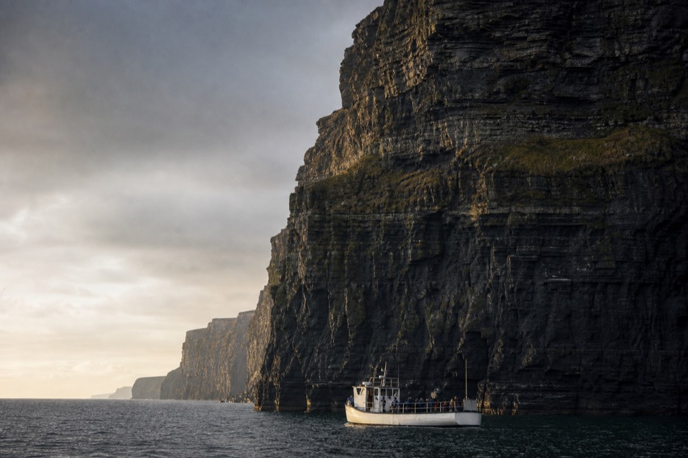 A small white passenger boat sitting low at the foot of the Cliffs of Moher, the dark layered rock wall towering hundreds of feet above it under a grey sky.