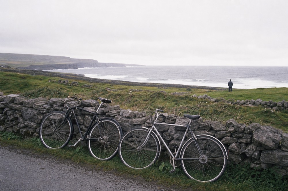 Bicycles leaning against a low stone wall on a green Aran island, the Atlantic and a distant line of cliffs visible beyond the fields under a bright sky.