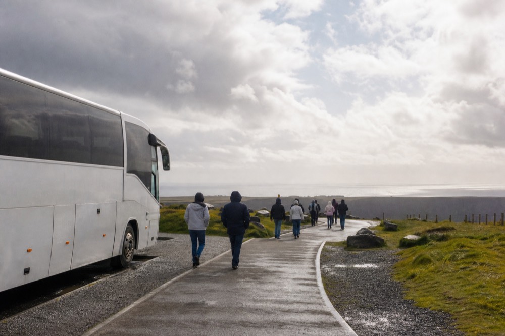 A tour coach parked at a green Irish coastal car park, passengers setting off along a paved path toward the Cliffs of Moher under a broad, breezy sky.