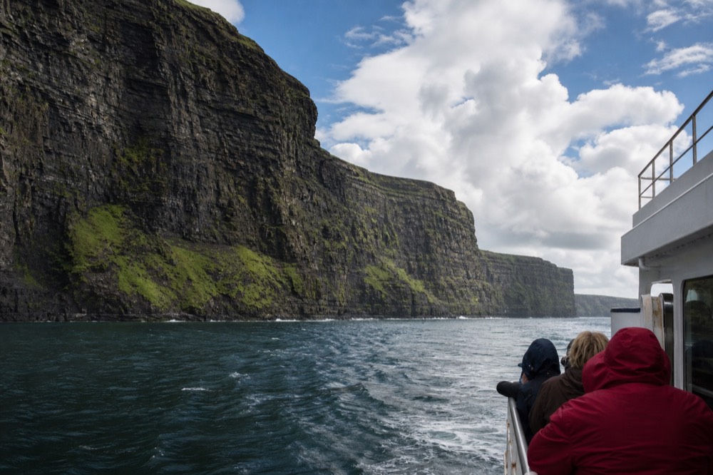 A cliff-cruise boat passing beneath the full height of the Cliffs of Moher, the horizontal bands of dark rock streaked with green rising steeply above the deck.