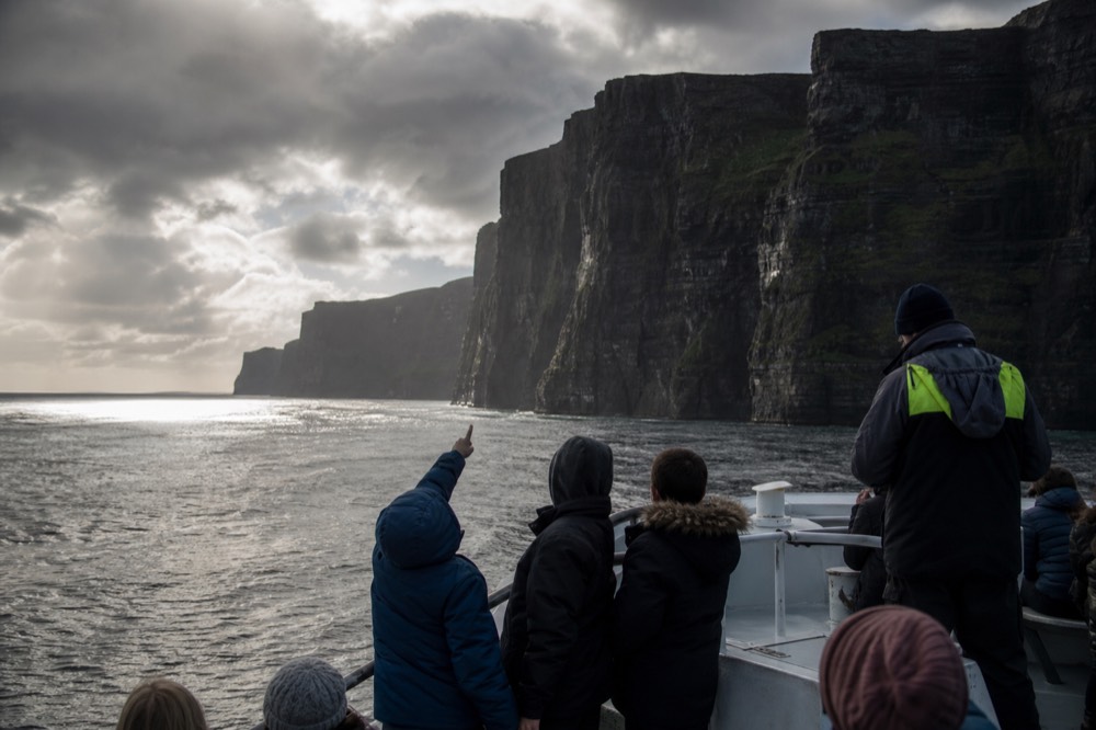 A family on the open deck of a cliff-cruise boat, children pointing up at the towering Cliffs of Moher, the crew nearby and calm water around the hull.
