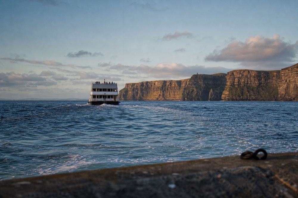 The Cliffs of Moher glowing amber in low evening light behind a returning cruise boat, its wake spreading across a calm, gently rolling sea.