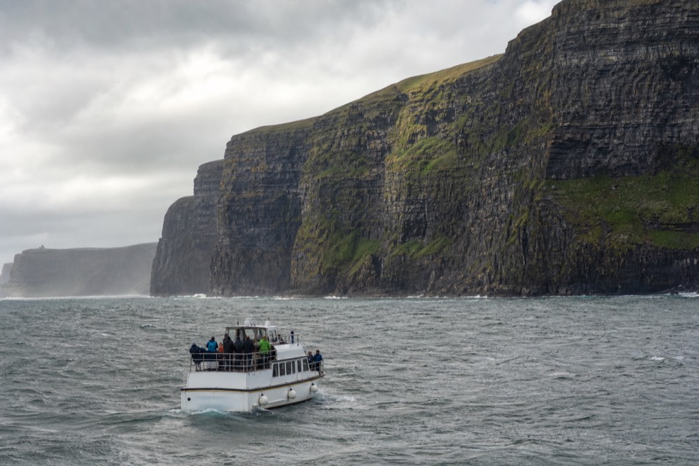 A small white cliff-cruise boat riding low over a grey rolling Atlantic swell directly below the towering Cliffs of Moher, passengers gripping the rail.