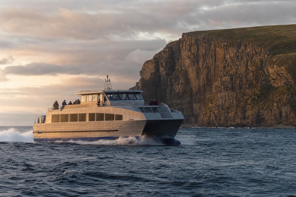 A large modern aluminium passenger ferry with a high enclosed cabin cutting steadily through open water off the County Clare coast, spray low at the bow.