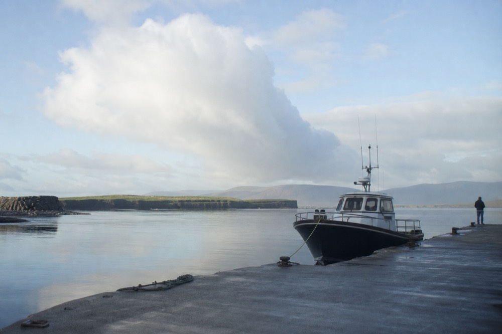 Doolin Pier at first light on a still morning, the harbour water glassy and calm, a tour boat moored ready with the Clare hills soft behind.