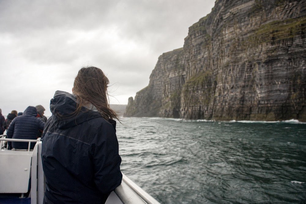 A passenger in a waterproof jacket standing at the open rail of a tour boat, looking steadily out at the horizon and the cliff wall, wind in their hair.