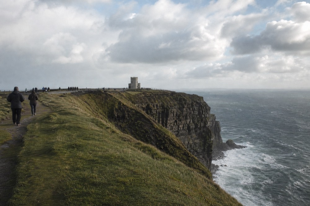 Walkers on the grassy clifftop path at the top of the Cliffs of Moher on a breezy day, the safe fenced viewpoint and O'Brien's Tower in the distance.