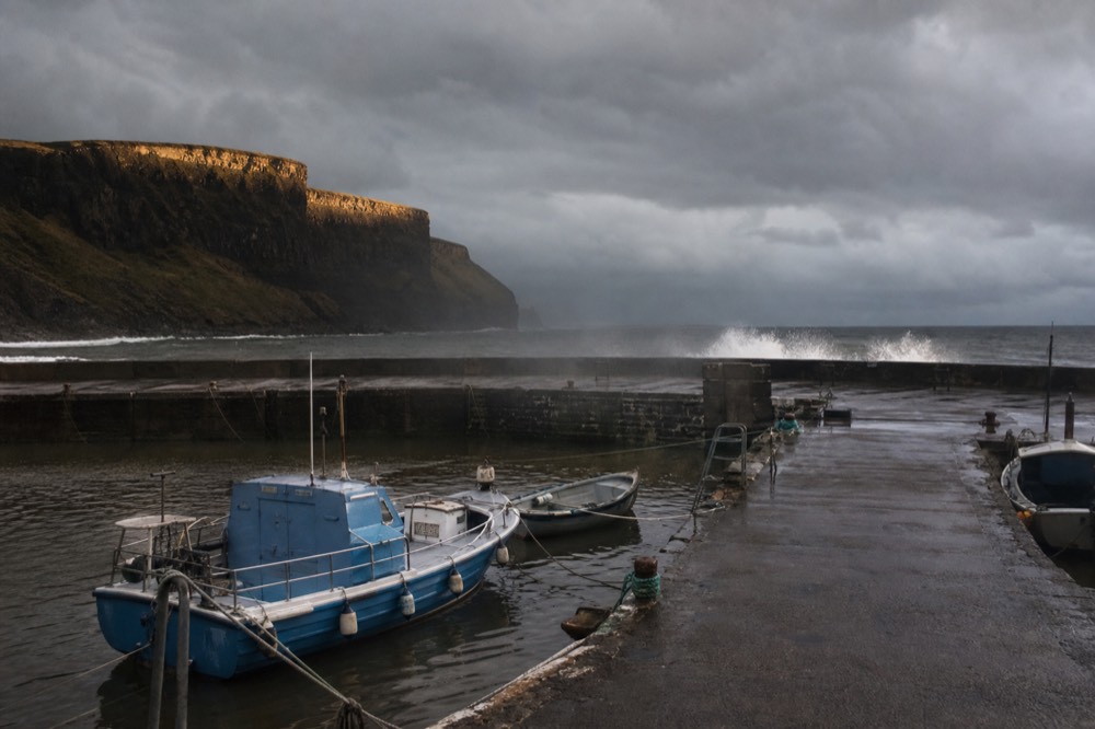 An empty Doolin Pier on a stormy grey day, boats tied up and idle, wind driving spray over the harbour wall under a heavy sky.