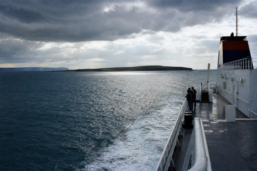 A large Aran Islands ferry crossing open blue water on a bright day, the low green profile of the islands ahead and the County Clare coast behind.