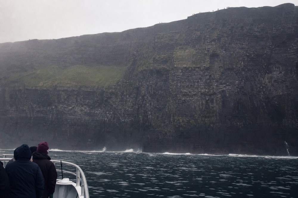 The Cliffs of Moher seen from a boat at their base, the vast dark wall filling the frame above a band of white surf, tiny figures just visible on the clifftop far above.