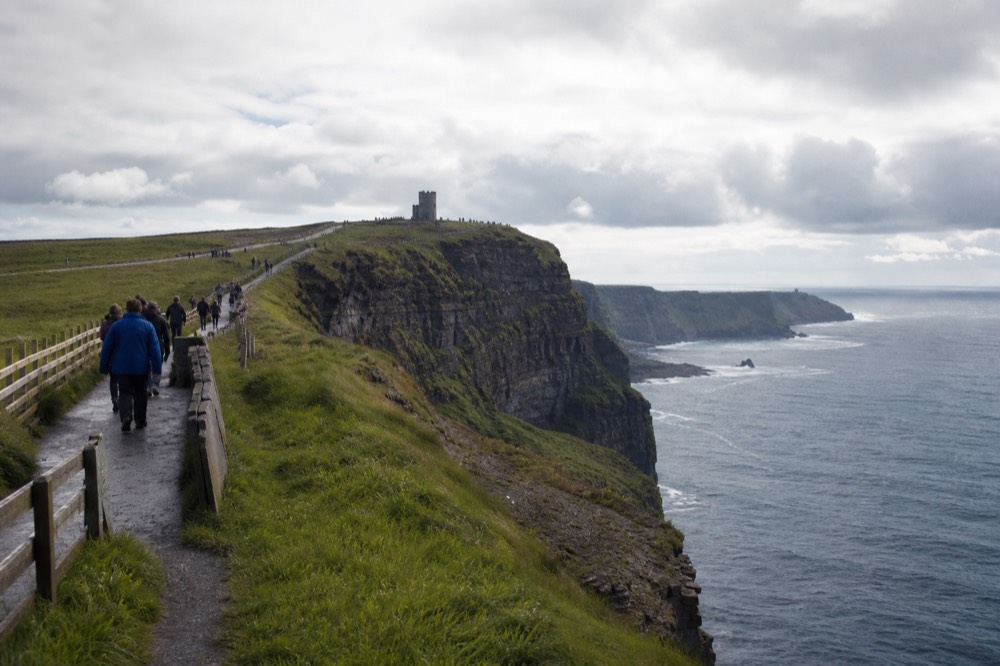 Walkers on the fenced clifftop path at the Cliffs of Moher on a bright day, O'Brien's Tower on the high point ahead, the coastline curving away below them.