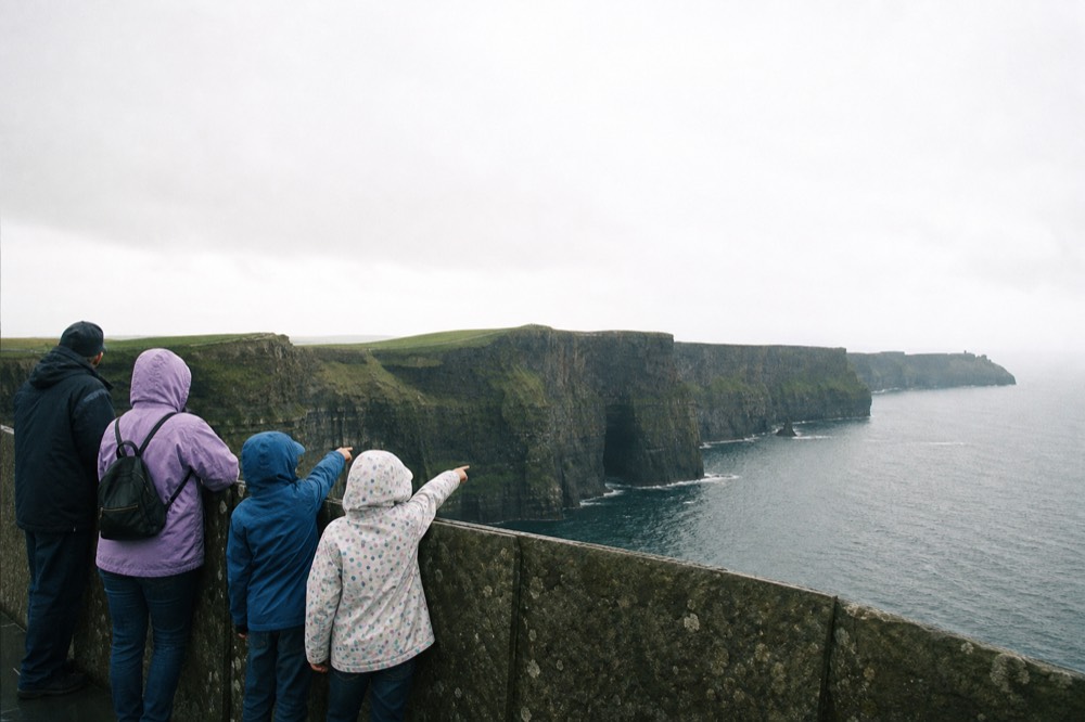 A family on the clifftop viewing area at the Cliffs of Moher, children at the safety wall pointing out to sea, the cliffs stretching away under a bright sky.