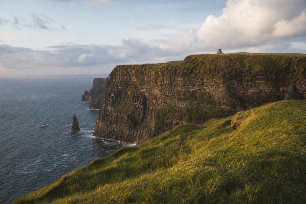The Cliffs of Moher at golden hour from the clifftop, warm light along the wall and O'Brien's Tower, a small boat visible far below at the base of the cliffs.