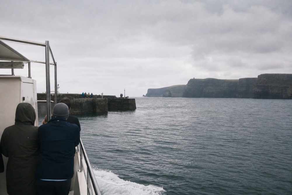 A cliff-cruise boat pulling away from Doolin Pier on a calm morning, the stone jetty behind and the open sea and cliffs ahead under soft light.