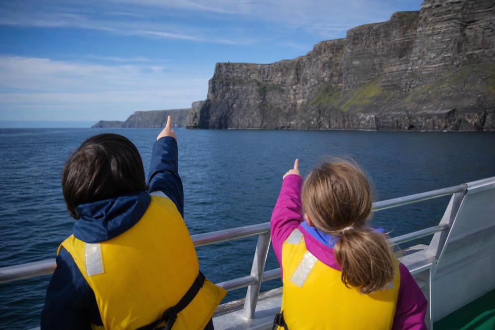 Two children in life jackets at the rail of a cliff-cruise boat, pointing up excitedly at the vast layered rock face of the Cliffs of Moher on a bright calm day.