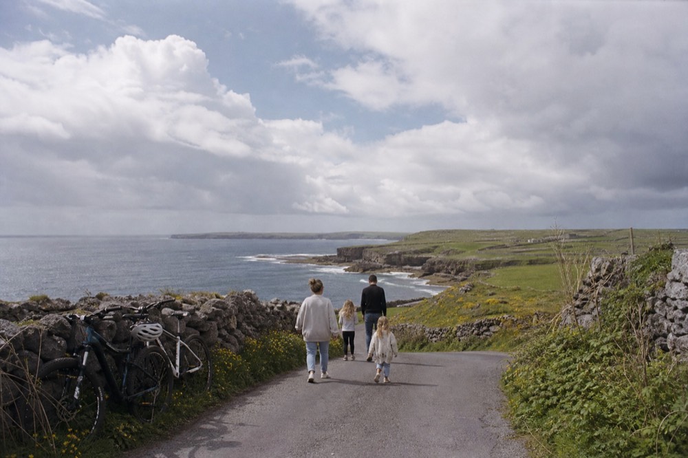 A family walking a low green Aran island lane on a bright day, bicycles leaning by a stone wall and the Atlantic wide behind them.