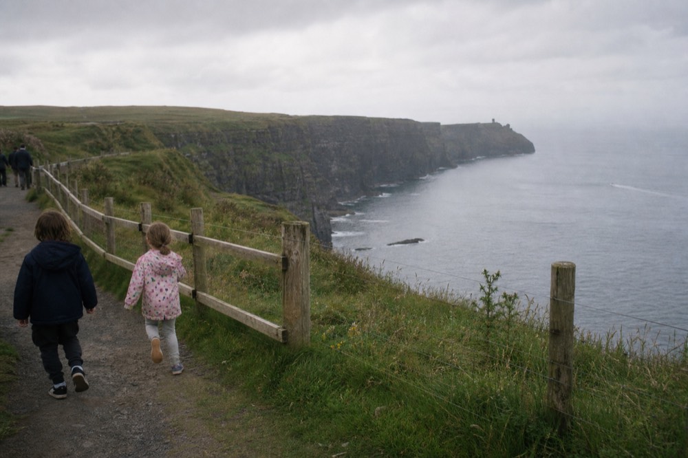 Children on the grassy clifftop path behind the safety fence at the Cliffs of Moher, the long green cliff line and O'Brien's Tower stretching away.
