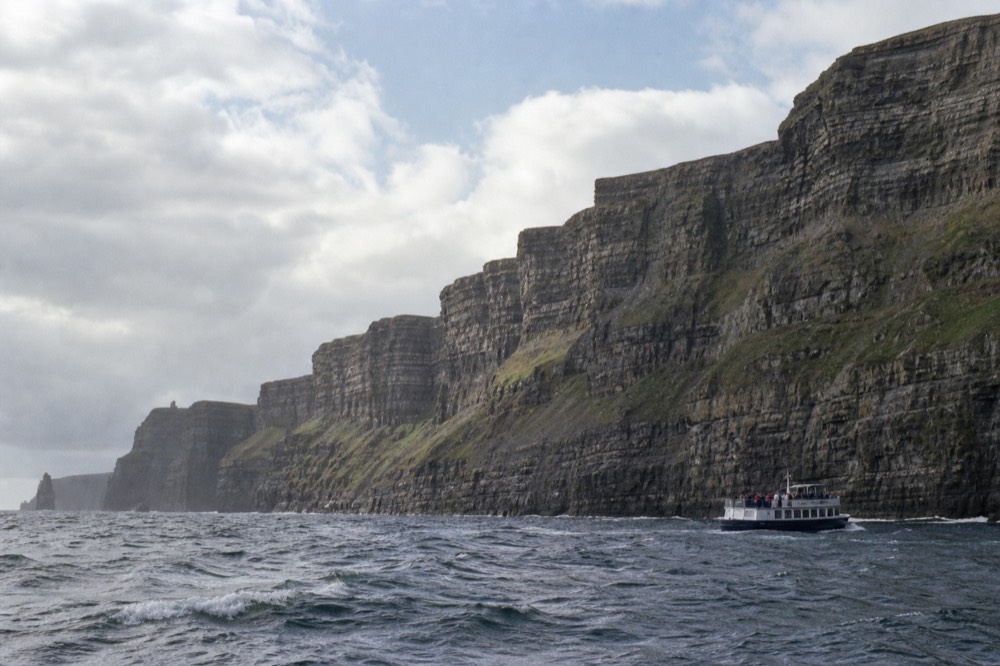 A cliff-cruise boat seen small at the foot of the Cliffs of Moher from sea level, the enormous rock wall dwarfing it under a soft bright sky.