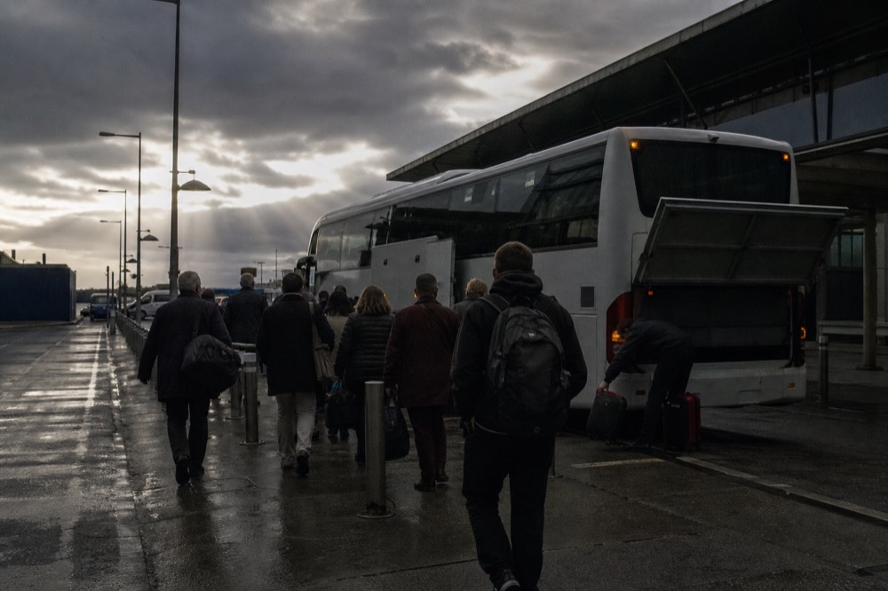 Passengers boarding a large tour coach outside a Dublin city terminus early in the morning, grey light and wet pavement, the driver loading bags below.