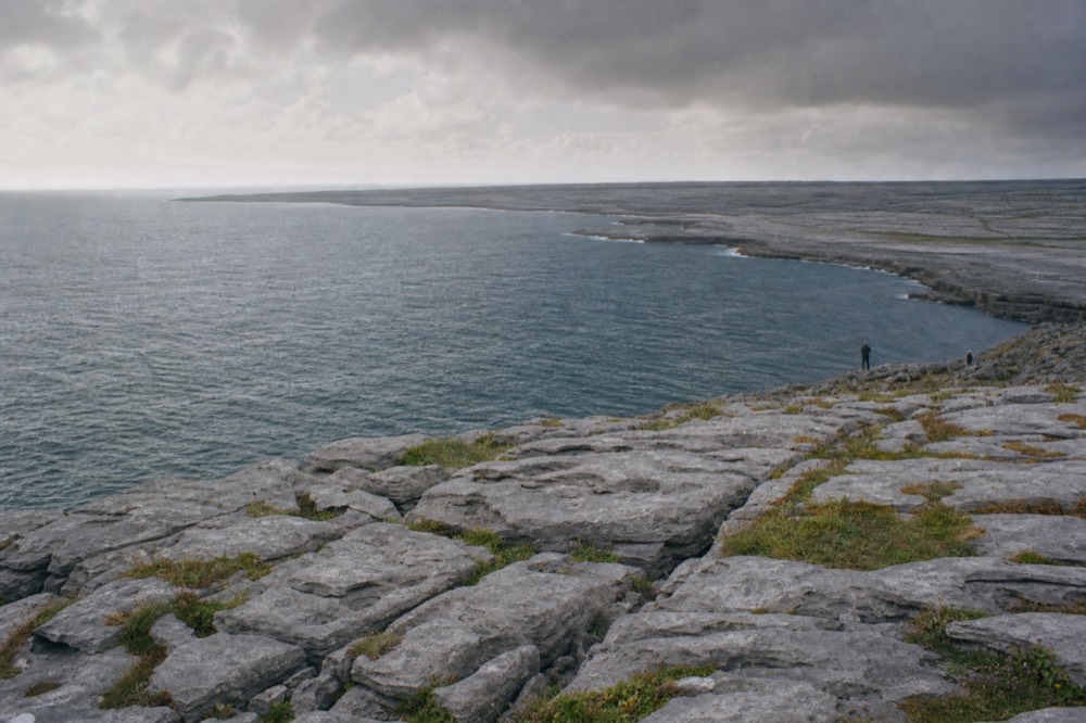 The grey limestone terraces of the Burren stretching to the horizon under a moody sky, bare rock pavement in the foreground with pockets of green between the cracks.