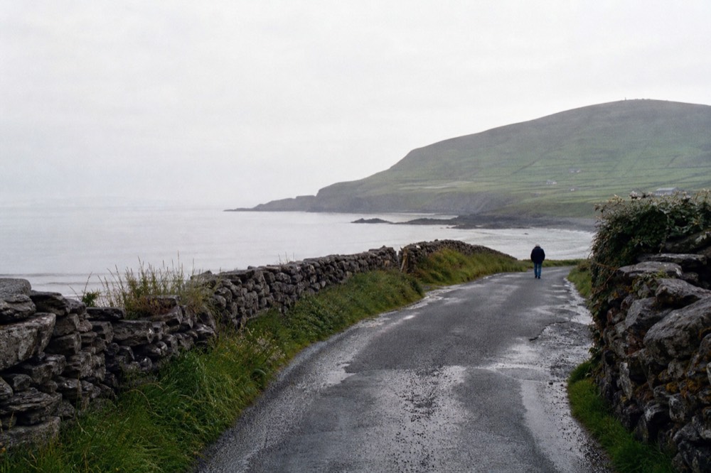 A narrow County Clare road winding between grey stone walls toward the coast, a glimpse of the Atlantic ahead and green hills rising on the right.