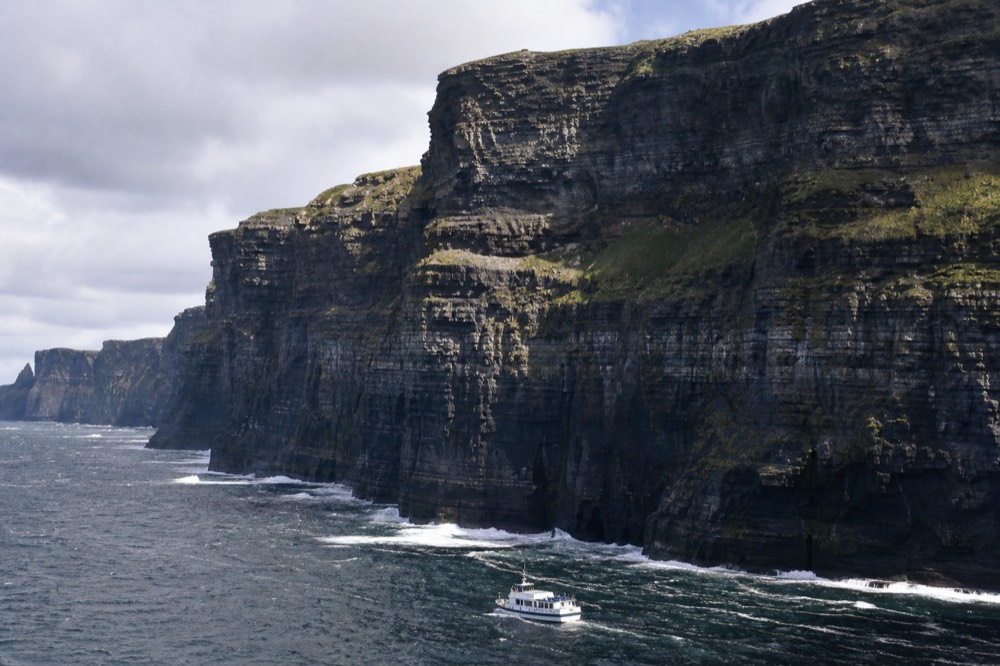 A cliff-cruise boat sitting small at the foot of the Cliffs of Moher, the dark layered rock wall filling the sky above a thin line of white surf.