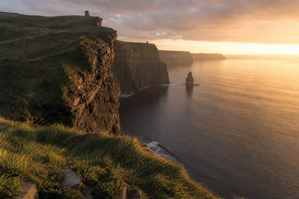 The Cliffs of Moher stretching south along the coast at golden hour, O'Brien's Tower small on the high clifftop, the sea calm and bright far below.