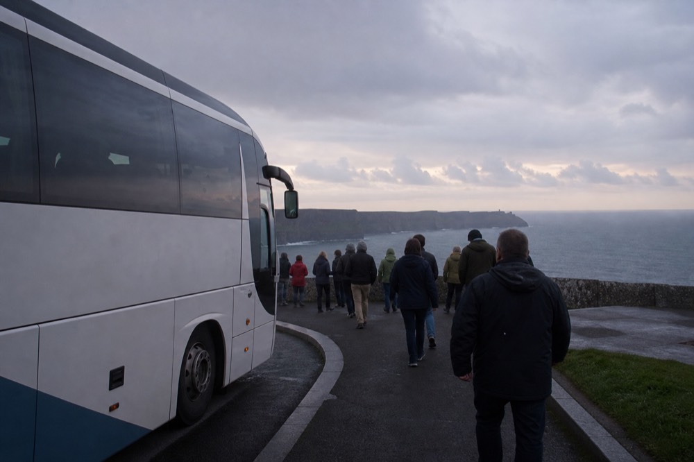A tour coach parked at a Clare coastal viewpoint at dusk, passengers stepping down toward a low wall with the darkening Atlantic and cliffs beyond.