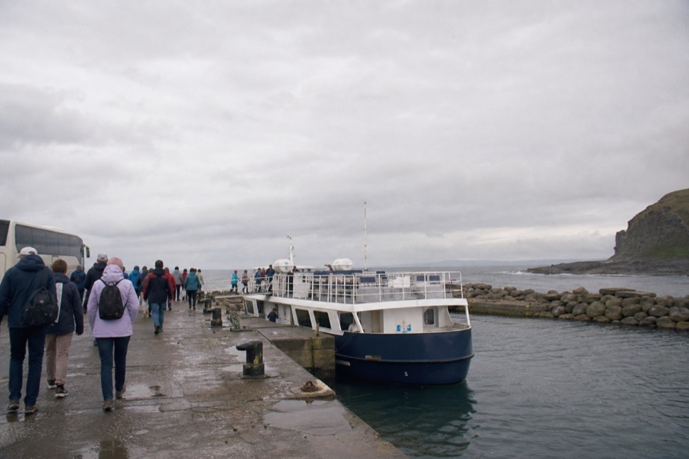 The small harbour at Doolin Pier with a cliff-cruise boat moored against the stone quay, tour passengers walking down from a parked coach to board.