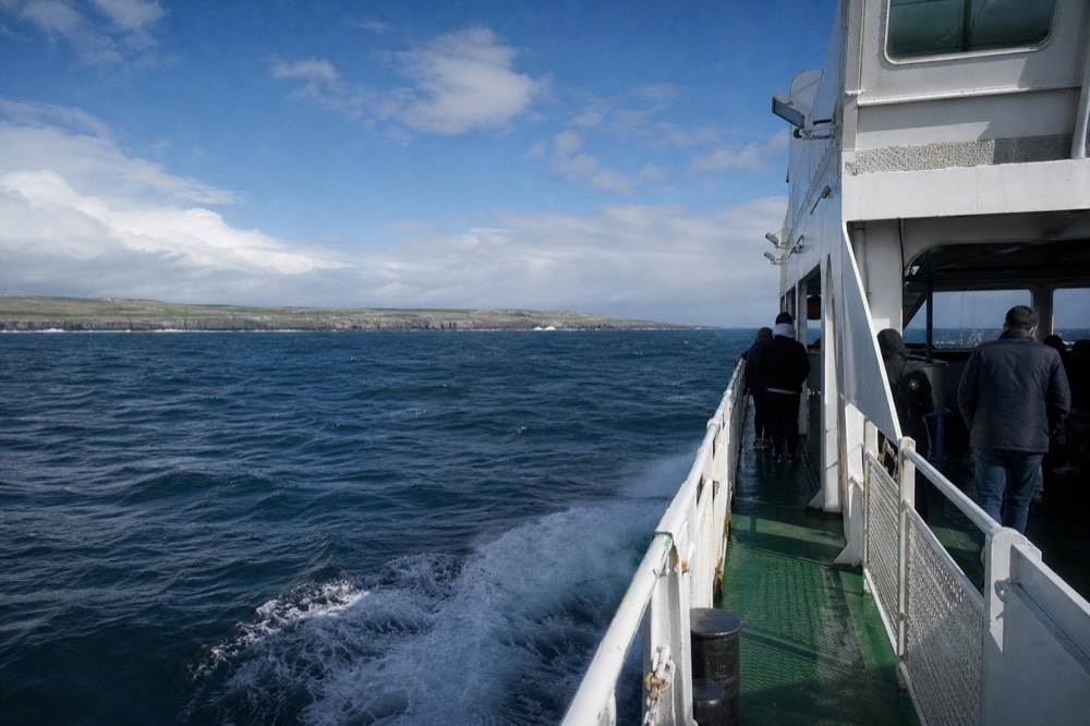 A ferry crossing open Atlantic water with the low green length of Inis Mor ahead, whitecaps on the swell and a bright break of sun over the island.