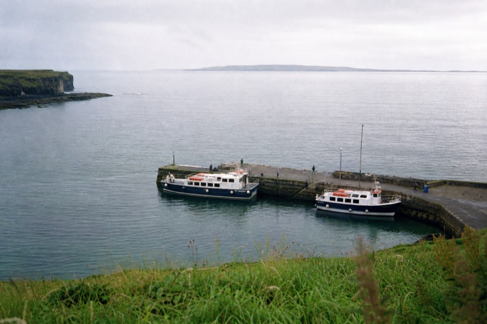 Doolin Pier from above, a short stone jetty with two cliff-cruise boats moored, green Clare hills behind and the Aran Islands low on the horizon.
