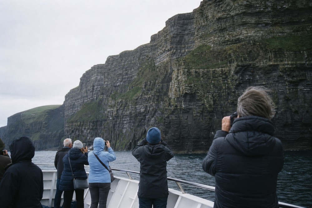 Passengers on the open deck of a cliff-cruise boat photographing the towering rock wall of the Cliffs of Moher rising close beside them under grey light.