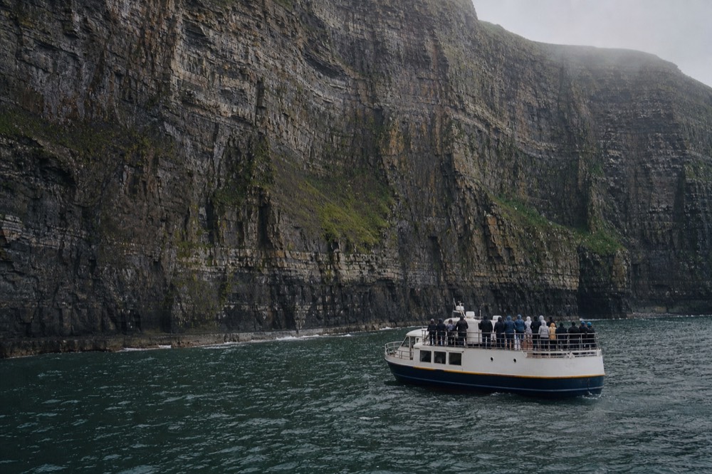 A cliff-cruise boat small against the base of the Cliffs of Moher, passengers along the rail tilting their heads back at the towering wall of layered rock above.