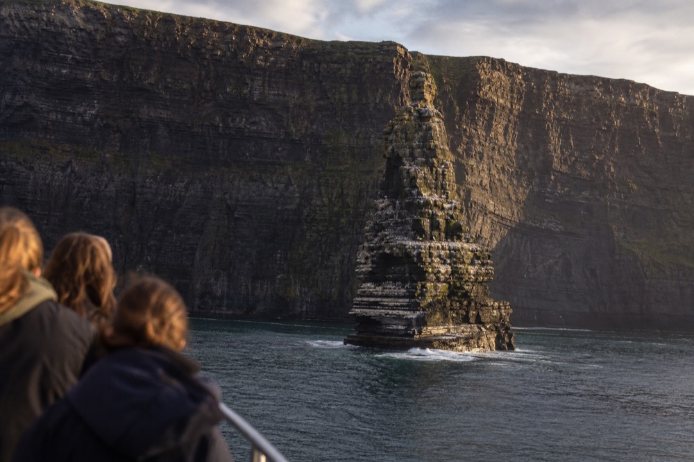 The An Branan Mor sea stack rising sheer from the ocean at the base of the Cliffs of Moher, its ledges white with nesting seabirds, the main cliff wall towering behind it.