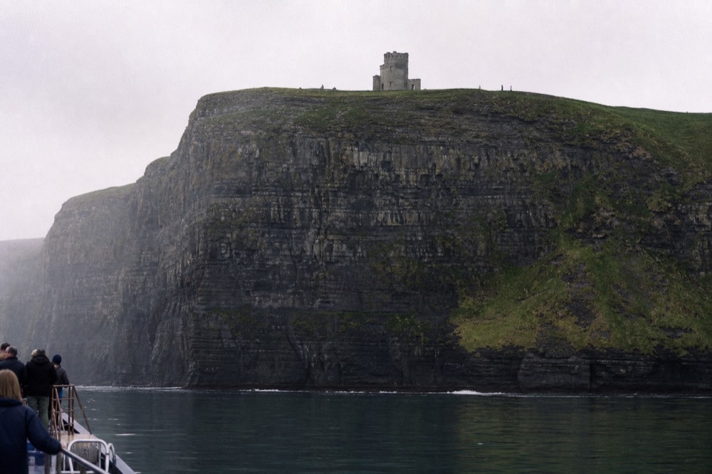 O'Brien's Tower, a small round stone tower, standing on the green clifftop high above, seen from a boat far below with the sheer cliff face falling away beneath it.
