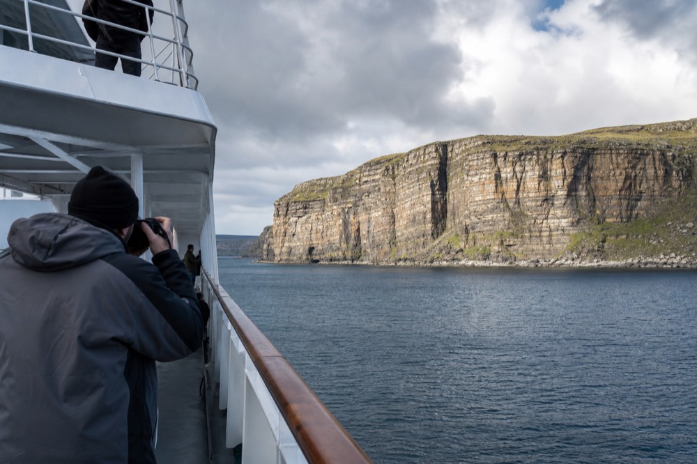 A photographer on the open deck of a cruise boat framing the sunlit cliff face at midday, the layered rock brightly lit and the sea calm and blue below.