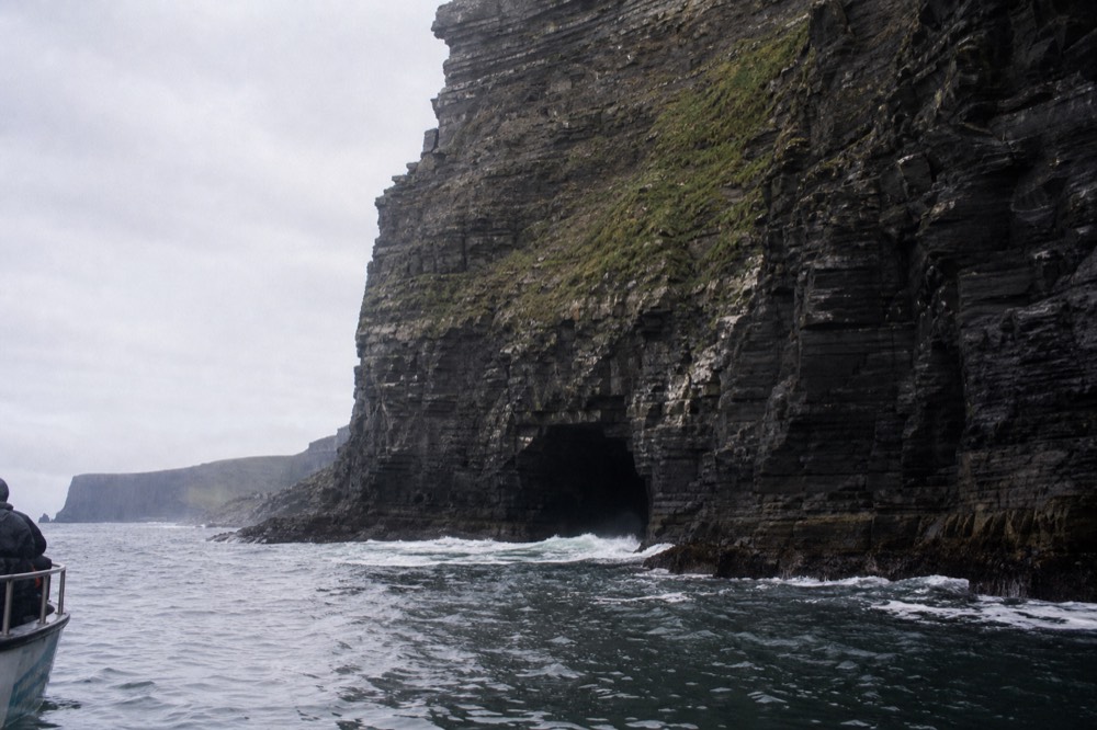 The dark mouth of the Harry Potter sea cave at the base of the Cliffs of Moher near Hag's Head, waves washing into the arch, the cliff towering above in shadow.