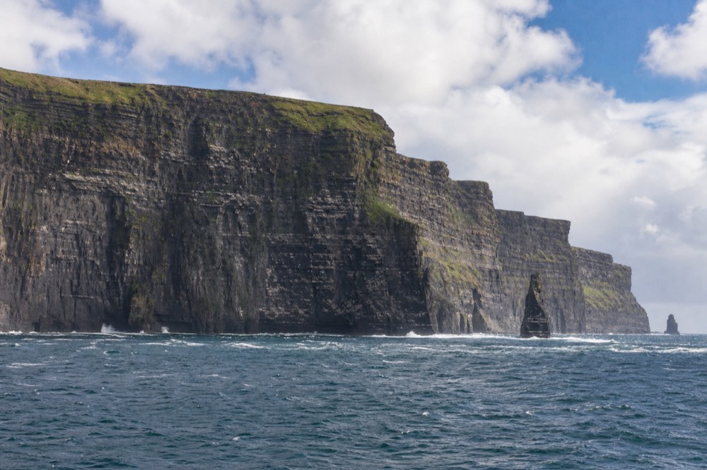 The full height of the Cliffs of Moher seen from sea level, horizontal bands of dark rock streaked with green, a thin line of white surf breaking at the base of the wall.