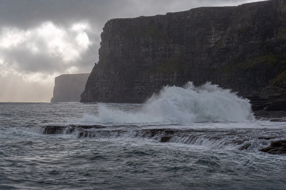 A large Atlantic wave breaking over the Aill na Searrach reef below the southern Cliffs of Moher, spray flung high, the dark cliff wall rising behind it.